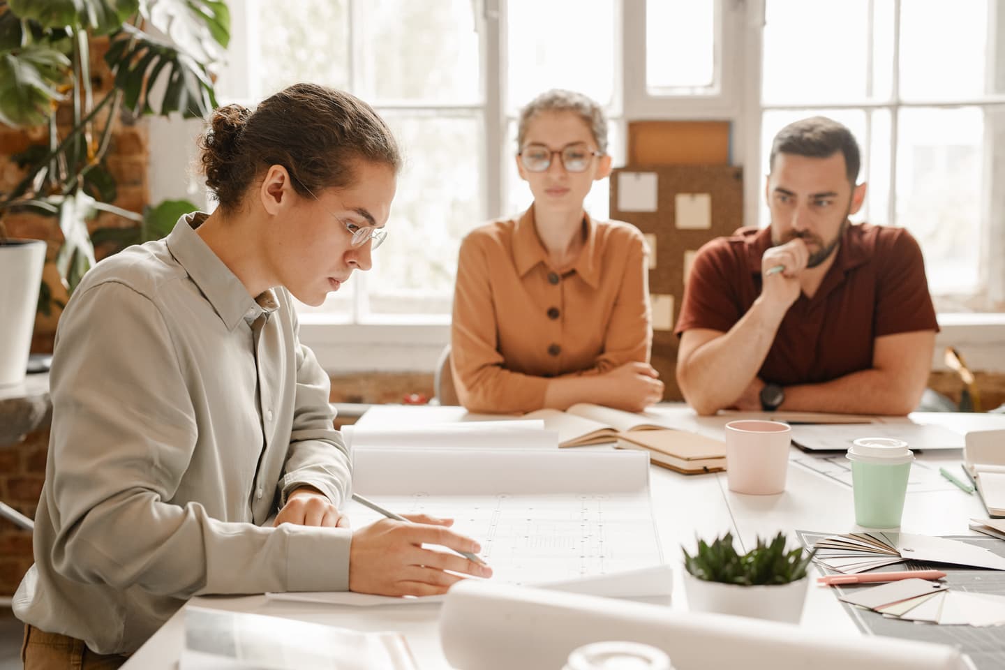 Three professionals in a bright office meeting reviewing documents with coffee cups and plants on the table.