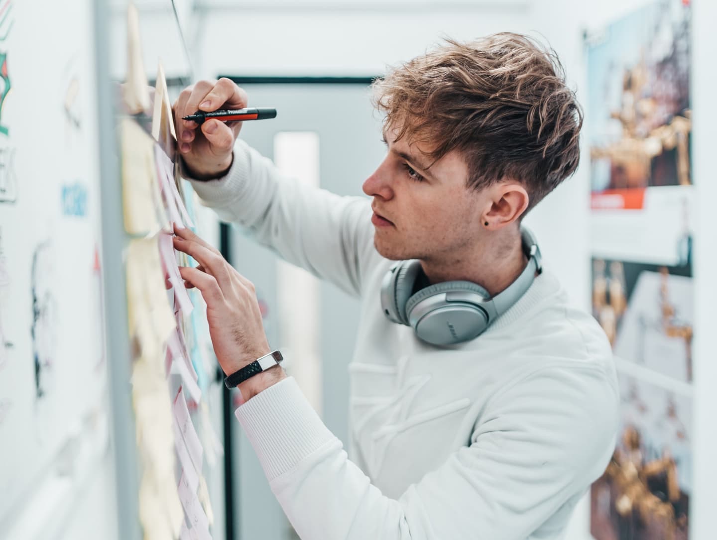Person in white jumper with headphones writing on wall notes, focused on creative planning in bright studio space.