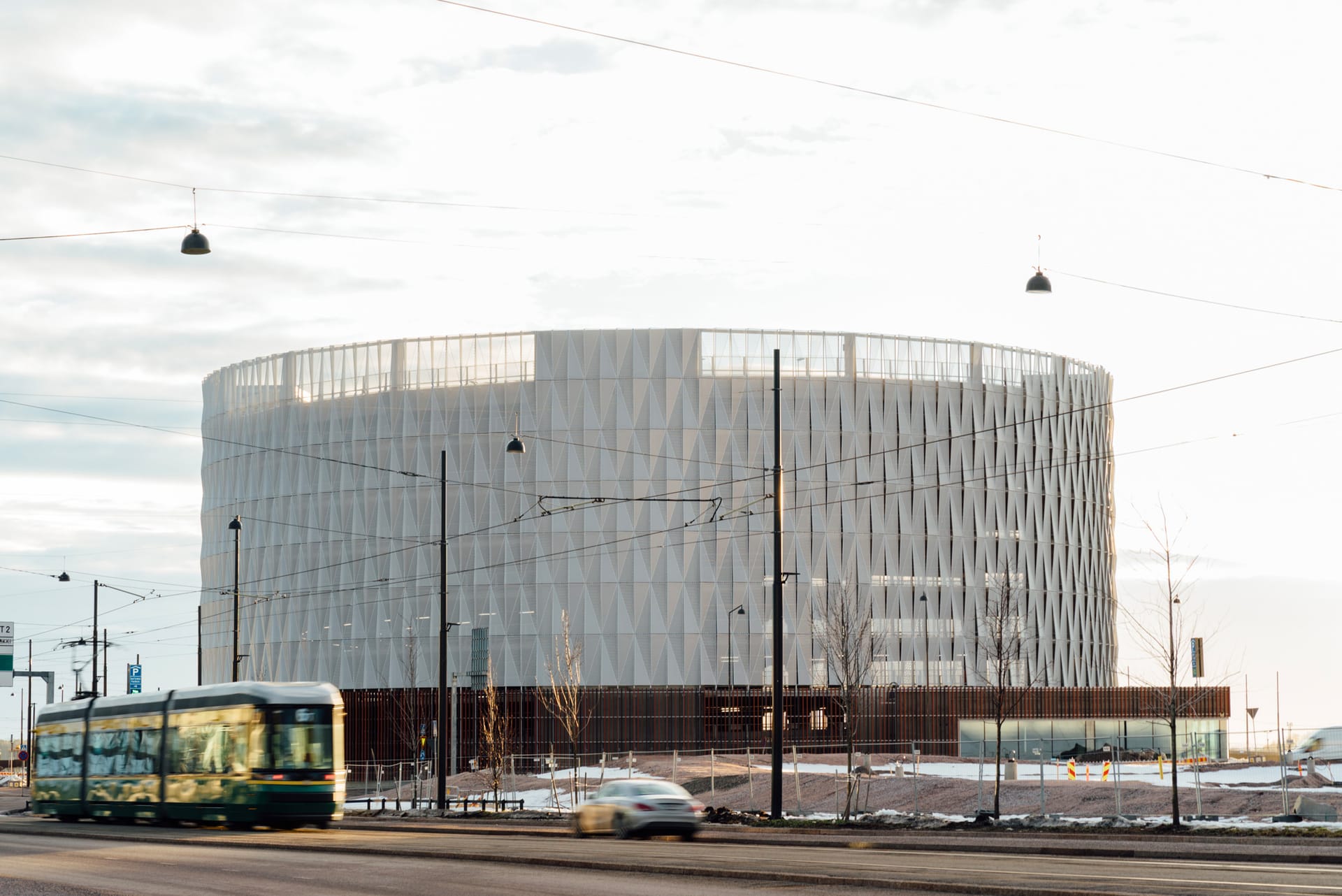 Modern circular building with geometric white façade, tram passing in foreground under cloudy sky.