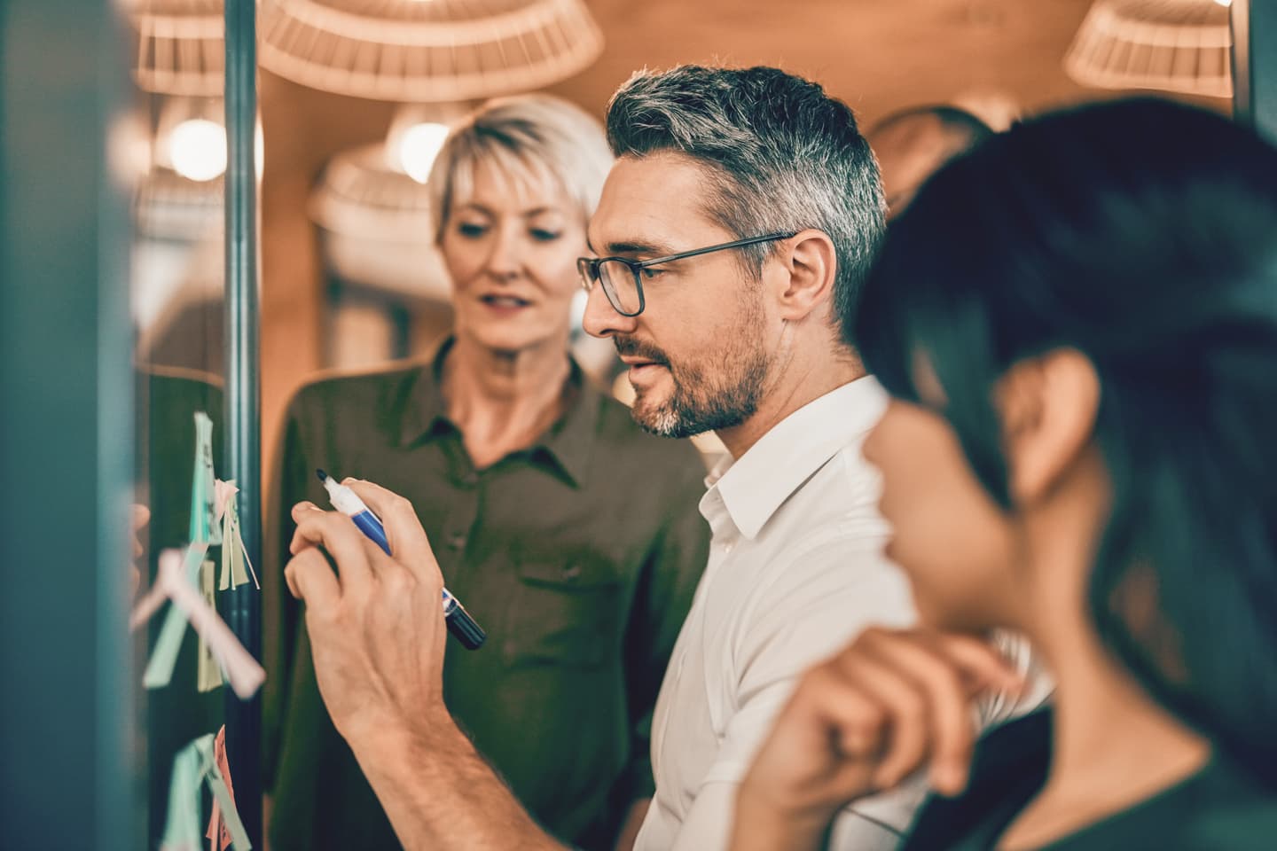 Two colleagues in an office setting reviewing notes on a glass board, with warm lighting and pendant lamps above.