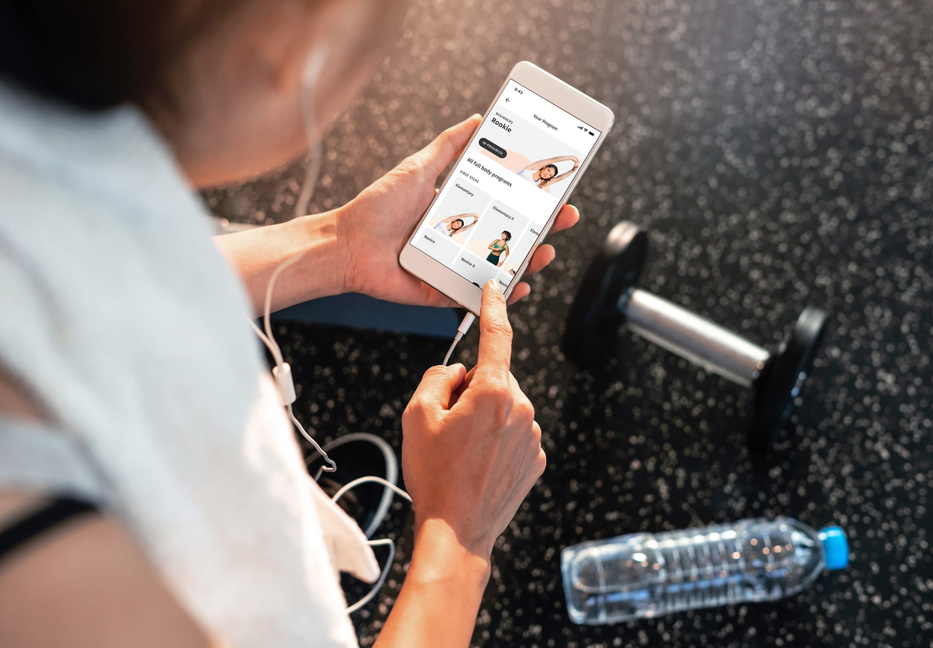 Person browsing fitness programs on smartphone at gym with water bottle and dumbbell nearby on dark floor.