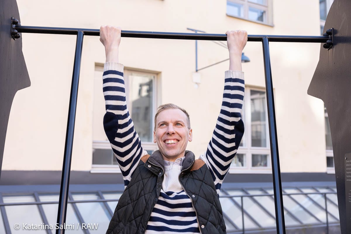 Person in striped jumper and black gilet doing pull-ups on outdoor bar, smiling against cream building backdrop.