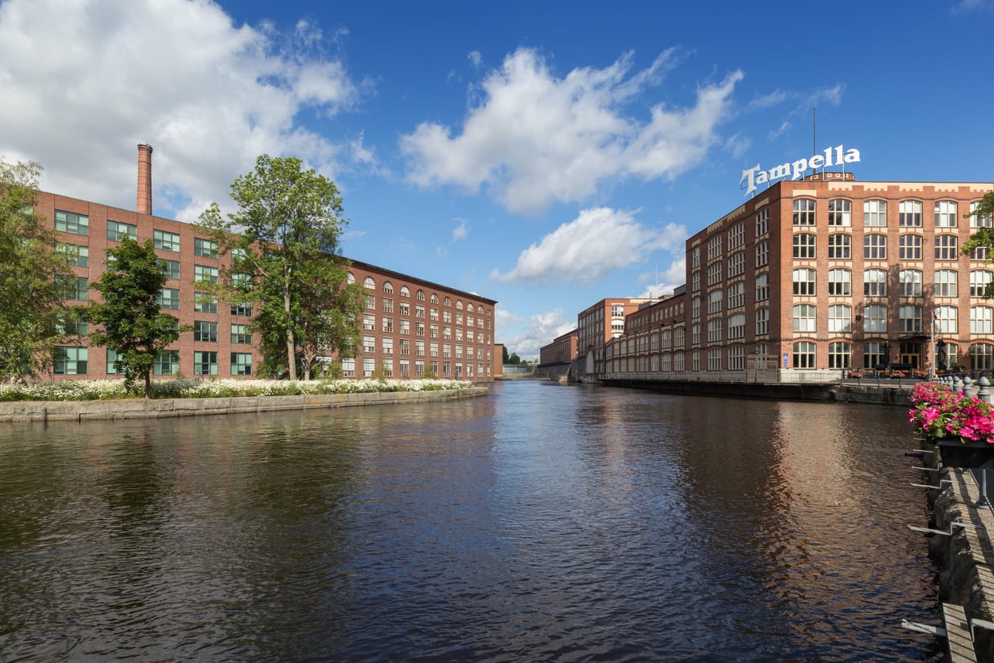Historic Tampella factory buildings along a canal in Tampere, Finland, with brick architecture under a blue sky.