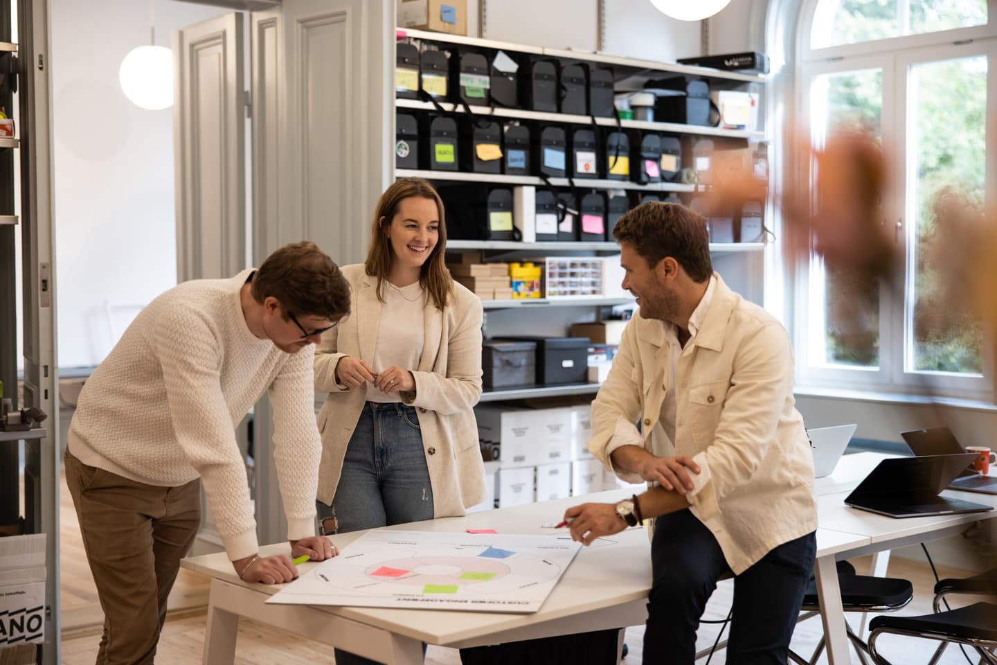 Three colleagues in light-coloured clothing collaborating over colourful charts in a bright office with organised shelving.