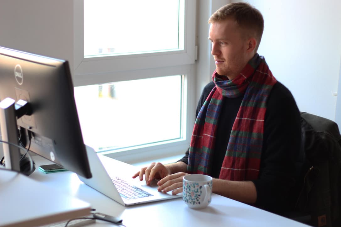Person in plaid scarf working on laptop at desk with coffee mug, seated by bright window.