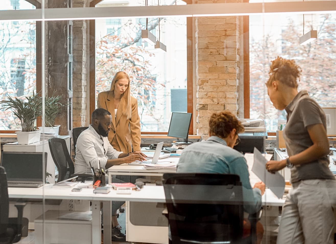Diverse team collaborating in modern office with exposed brick walls and large windows showing autumn trees outside.
