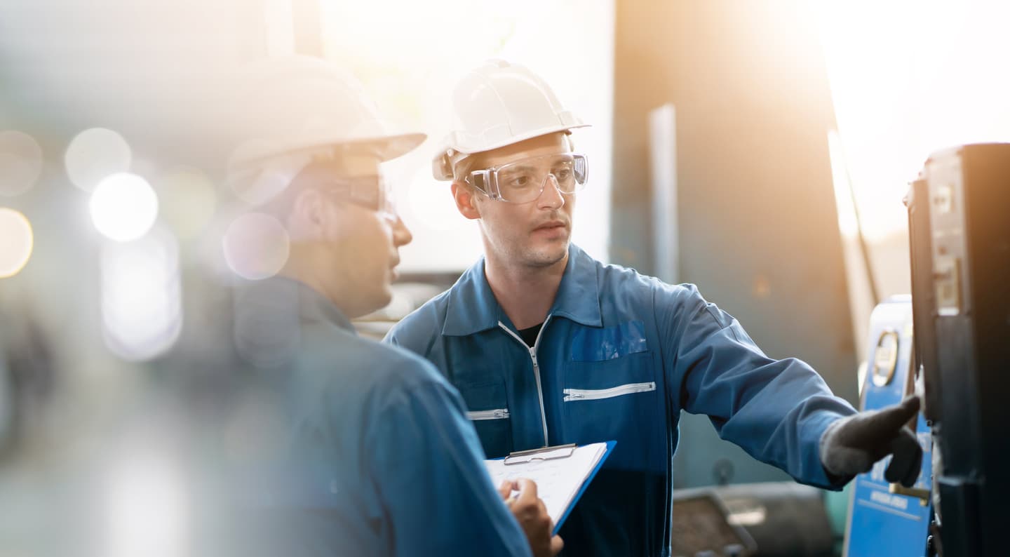 Industrial workers in blue uniforms and white hard hats examining equipment, one holding a clipboard.