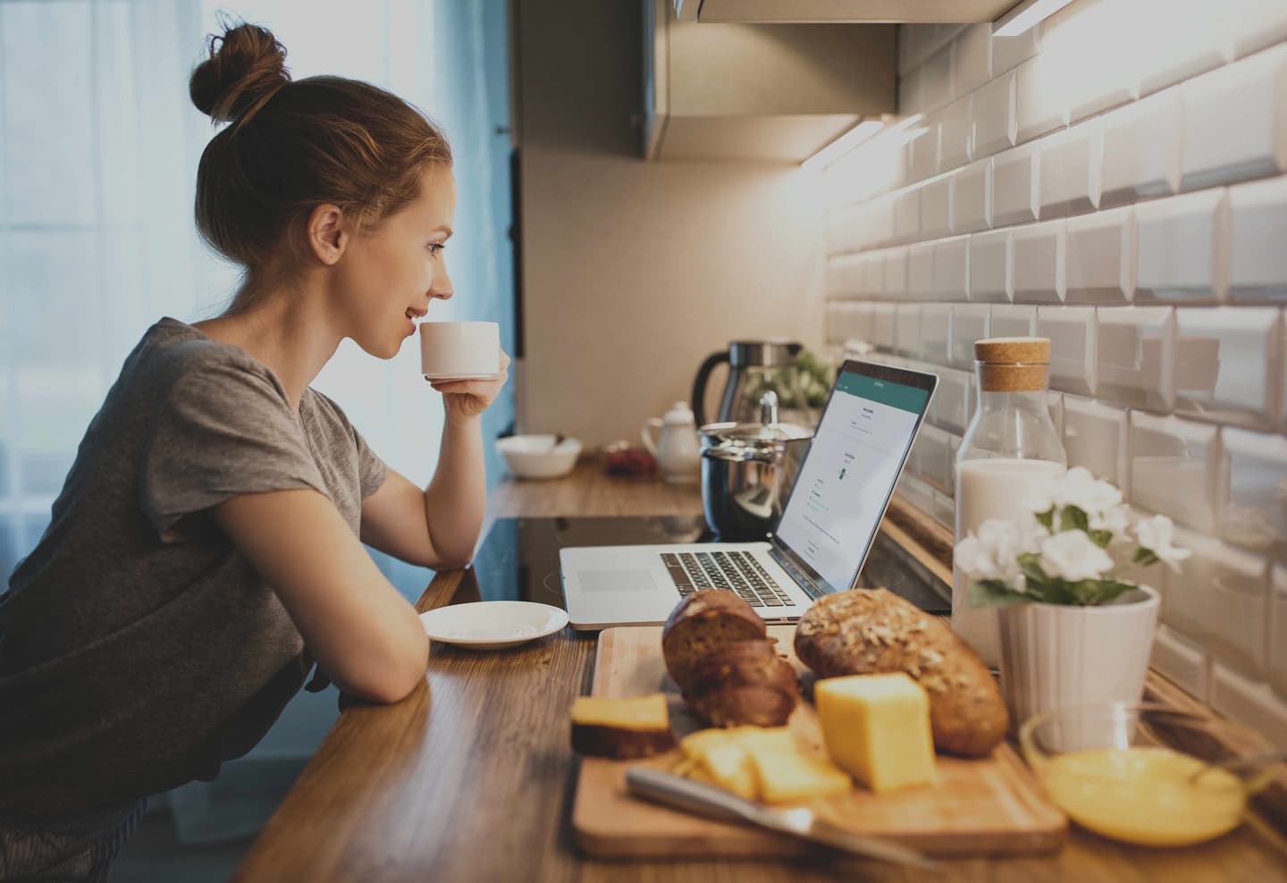 Person drinking coffee while using laptop in kitchen with bread and cheese on wooden board and white tiled backsplash.