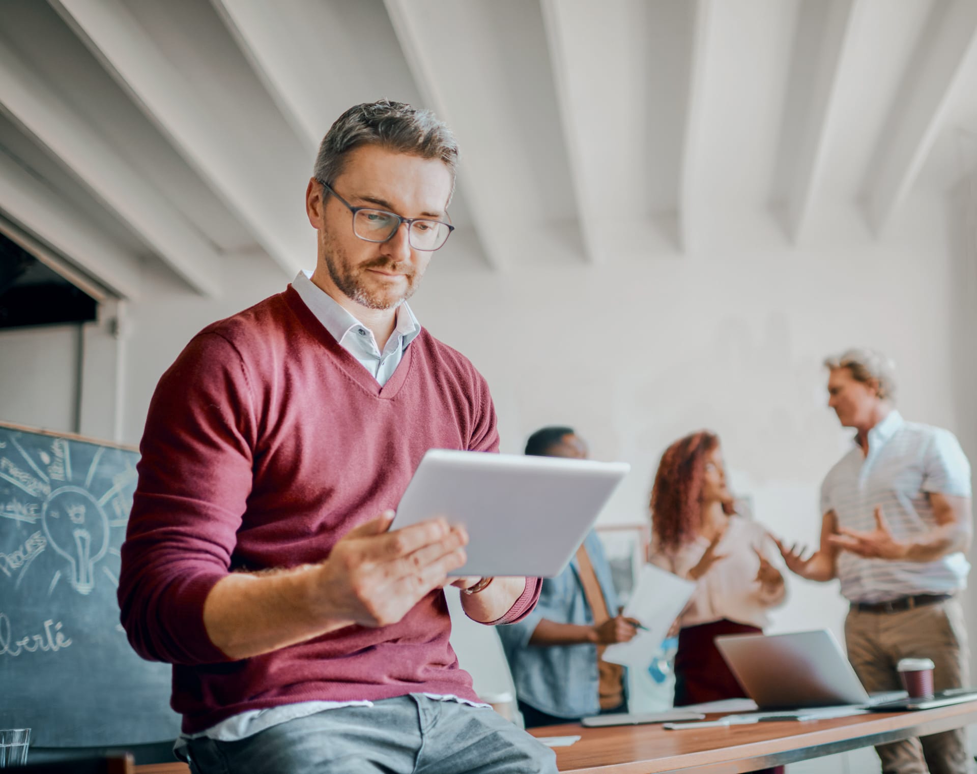 Person in burgundy jumper using tablet in modern office with colleagues discussing in background.