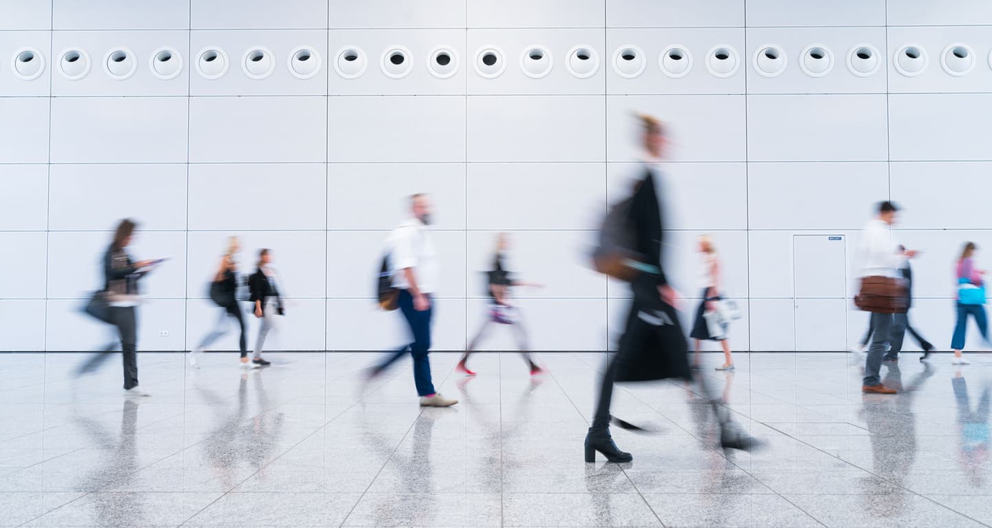 Blurred commuters walking past white tiled wall in modern building, creating sense of movement and urban hustle.