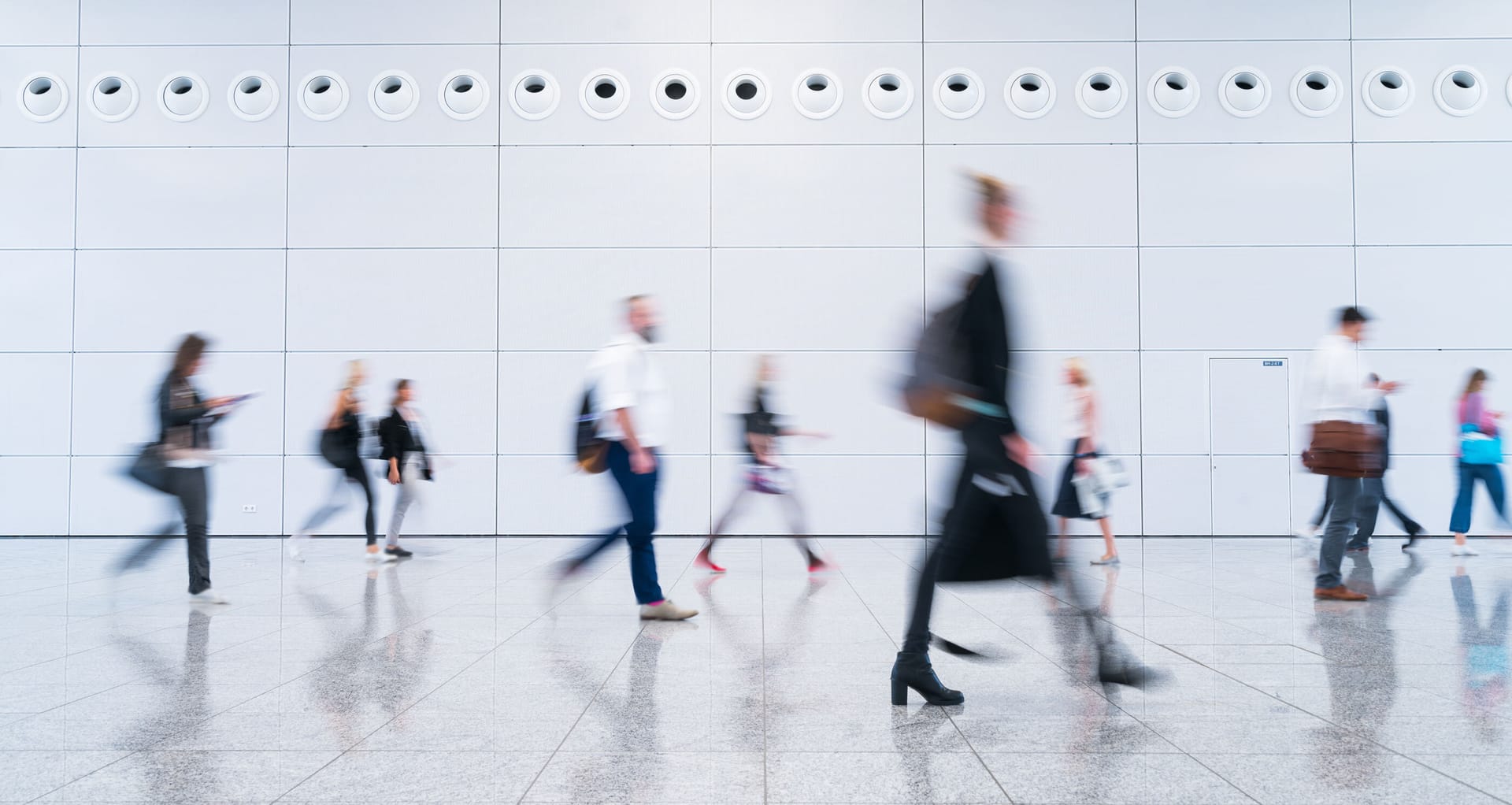Blurred commuters walking past white tiled wall in modern building, creating sense of movement and urban hustle.