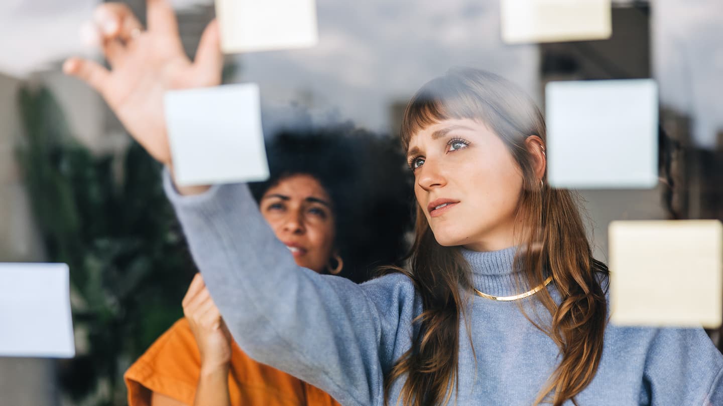 Two colleagues in blue and orange attire arranging sticky notes on a glass wall during a brainstorming session.