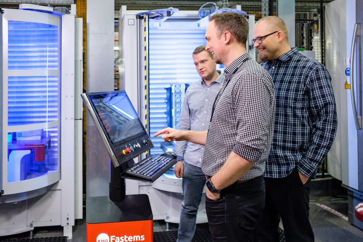 Three colleagues in a manufacturing facility operating a control panel with blue-lit industrial equipment in the background.