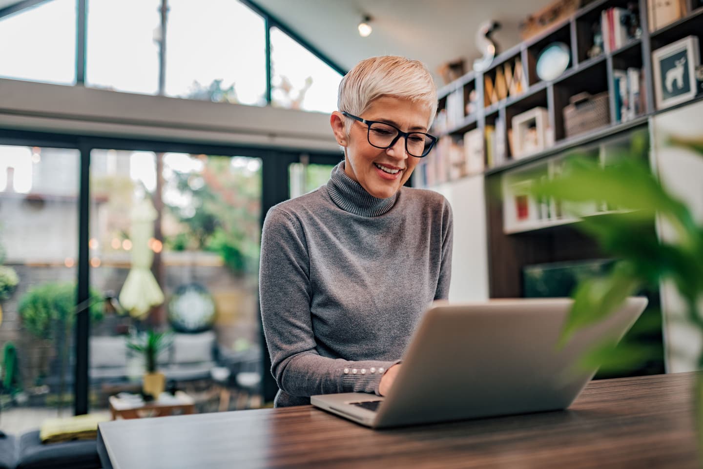 Person with short blonde hair and glasses smiling while using laptop in bright home office with bookshelves.