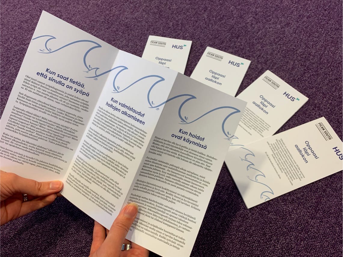 Hands holding Finnish healthcare brochures with blue wave designs on a purple carpet, HUS medical information pamphlets.