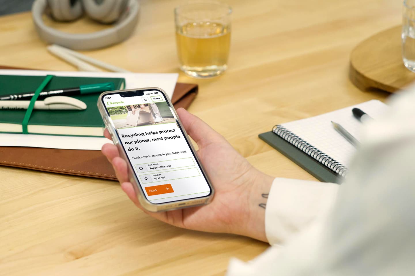 Hand holding phone displaying recycling app with search for paper coffee cups on wooden desk with notebook and drink.