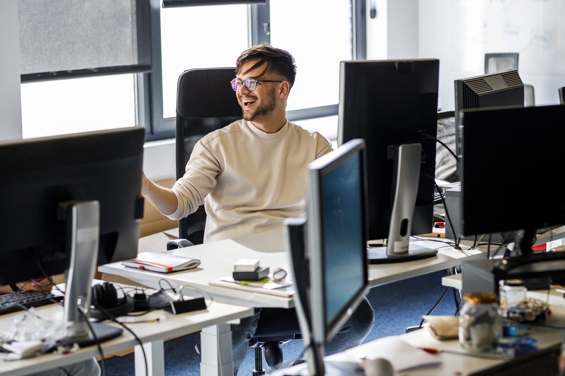 Person in cream jumper and glasses smiling at computer workstation in bright modern office with multiple monitors.