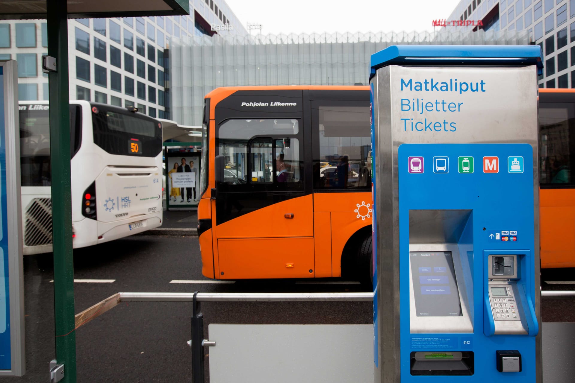 Blue ticket machine at bus station with orange and white buses in background, modern transport hub in Finland.