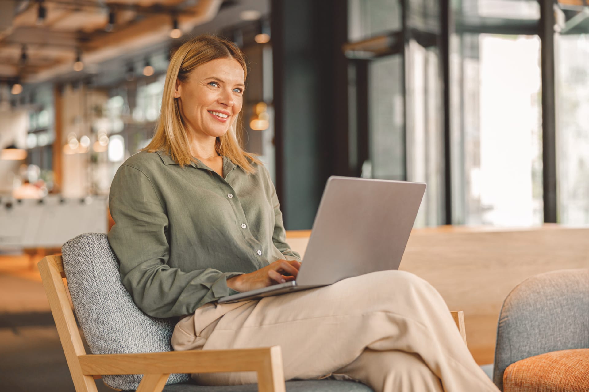 Woman in green shirt smiling while working on laptop in modern office space with large windows.
