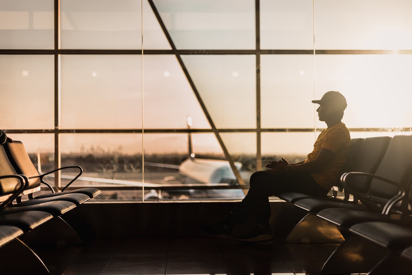 Silhouette of person waiting in airport terminal at sunset, with aircraft visible through large windows.