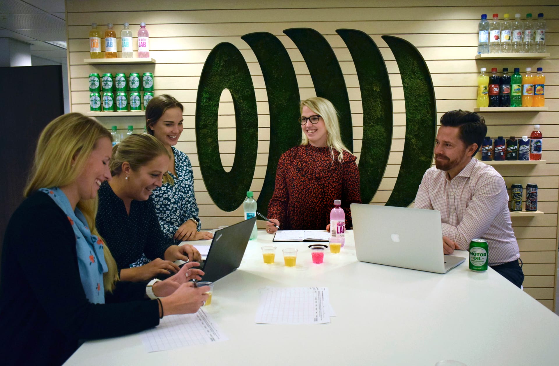 Group meeting in modern office with green moss logo wall, laptops on table, and beverage shelves displaying drinks.