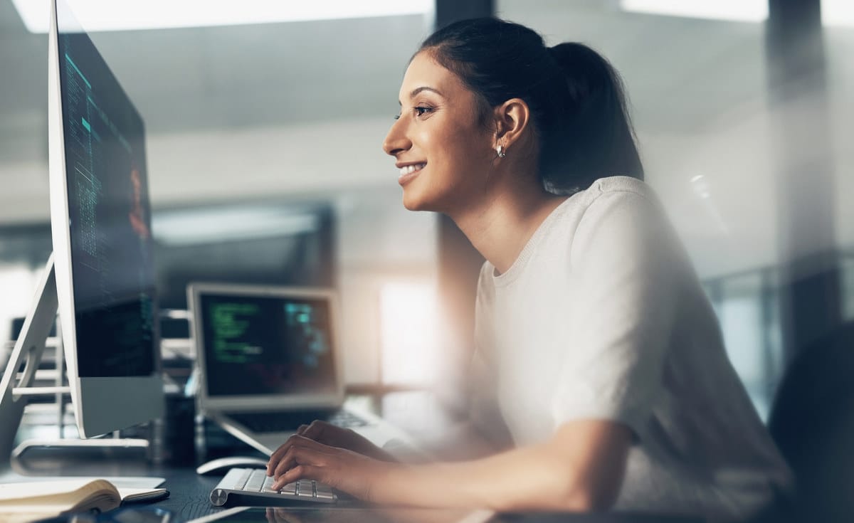 Person with ponytail smiling while working at computer in modern office with code visible on multiple screens.