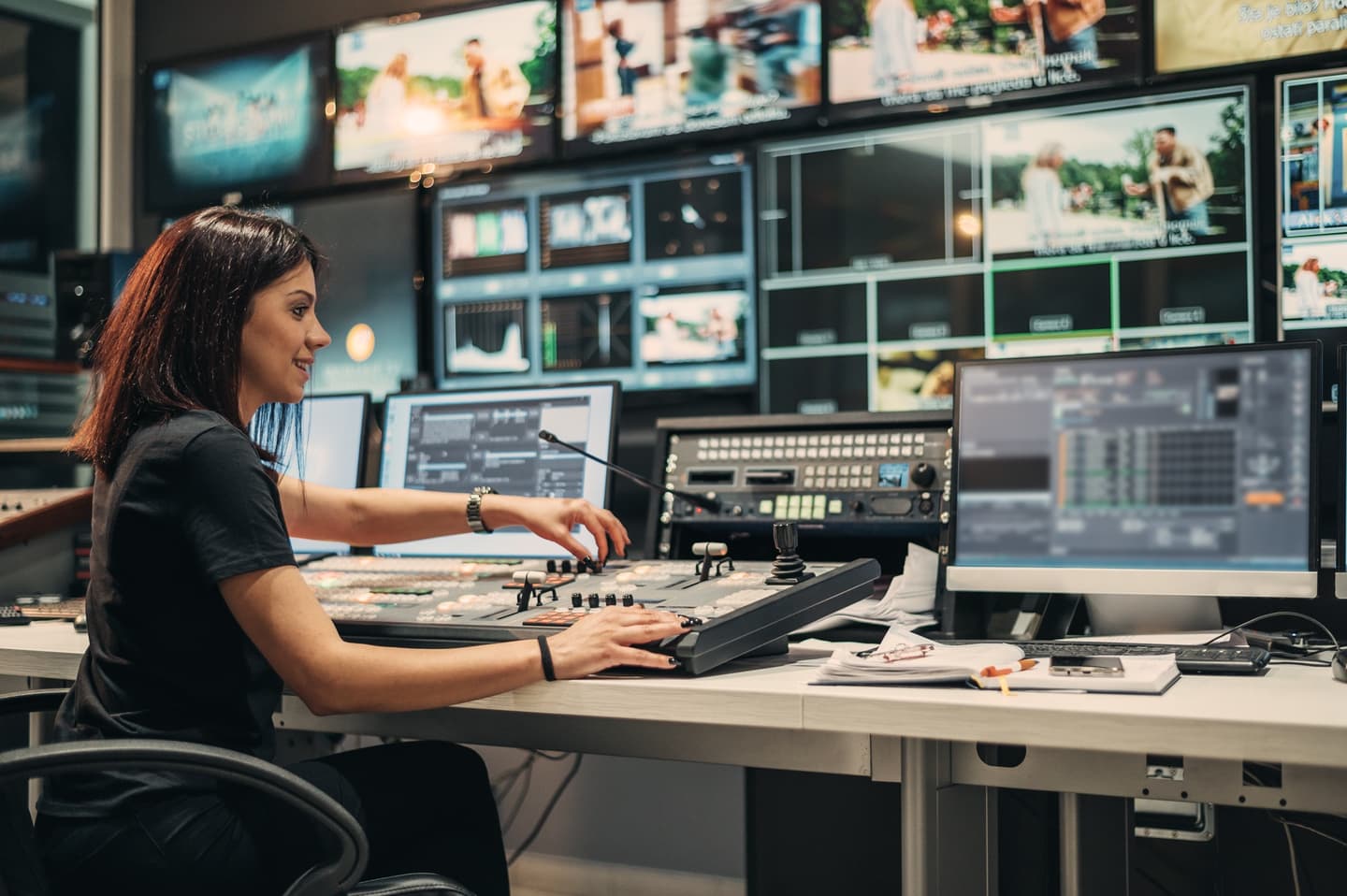 Person operating video editing equipment in a production control room with multiple monitors displaying content.