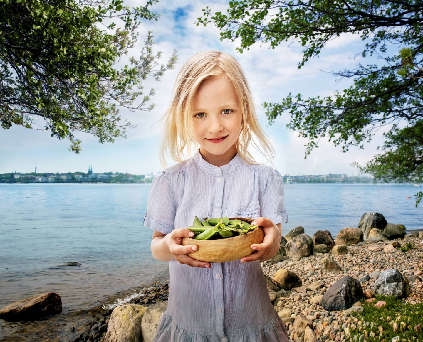 Child in light blue dress holding wooden bowl of greens by lakeside with rocky shore and city skyline in background.