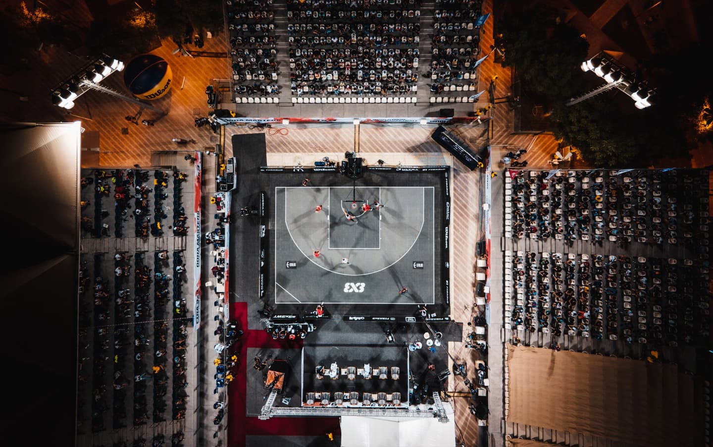 Aerial view of a 3x3 basketball court with players in action, surrounded by spectator seating under floodlights at night.