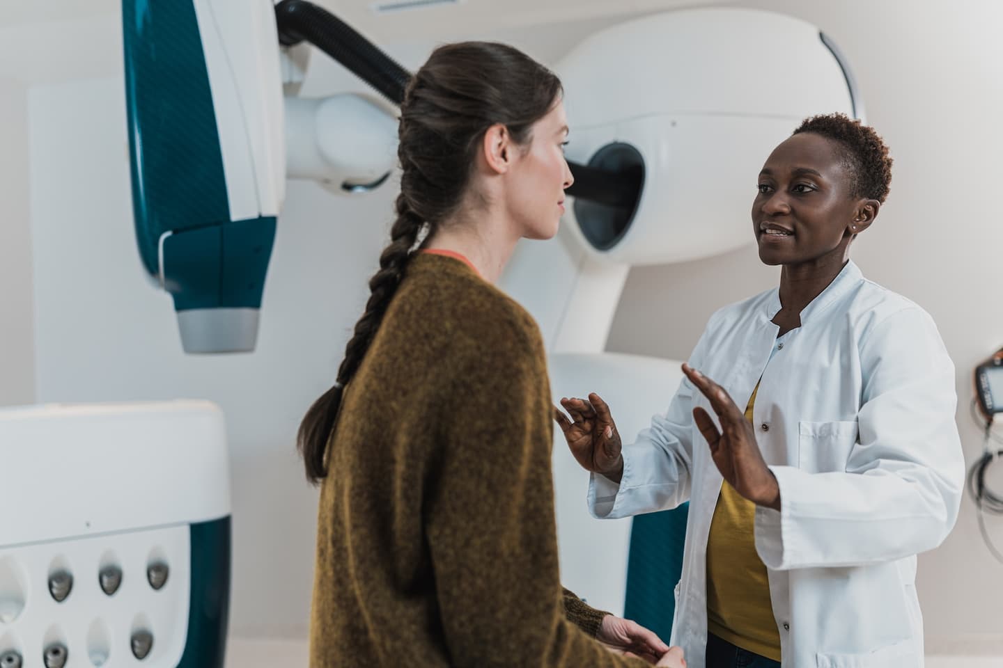Healthcare professional in white coat explaining procedure to patient in brown sweater near medical imaging equipment.