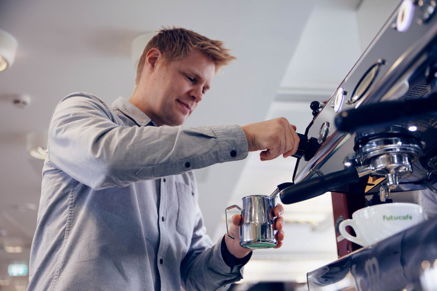 Person in light blue shirt operating an espresso machine, holding a metal milk jug in a bright café setting.