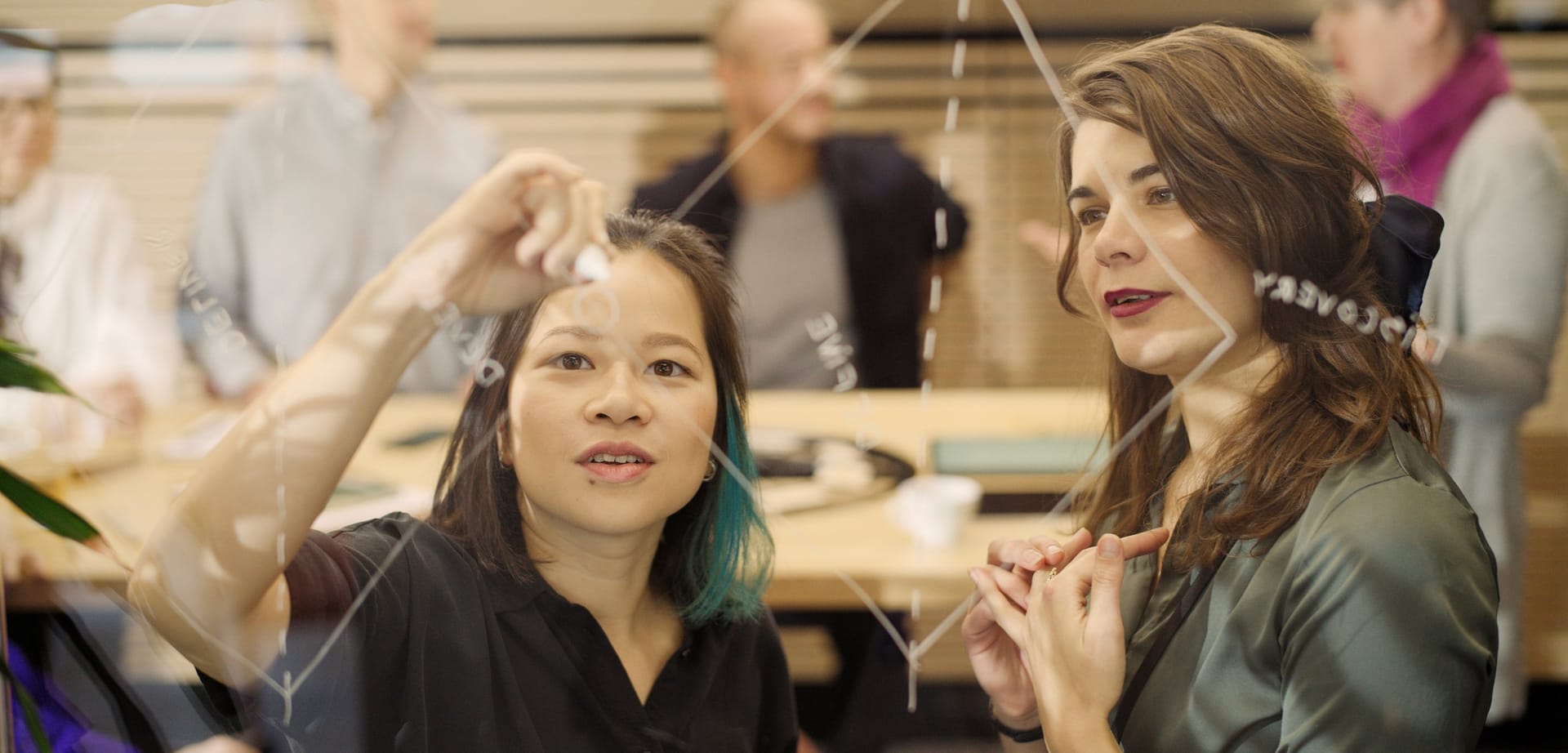 Two women collaborating on a transparent board, one pointing at notes while others work in the background.
