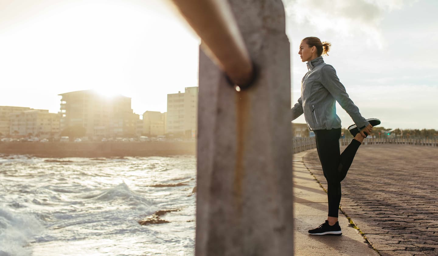 Runner stretching on waterfront promenade at sunset, wearing grey jacket and black leggings, city skyline in background.