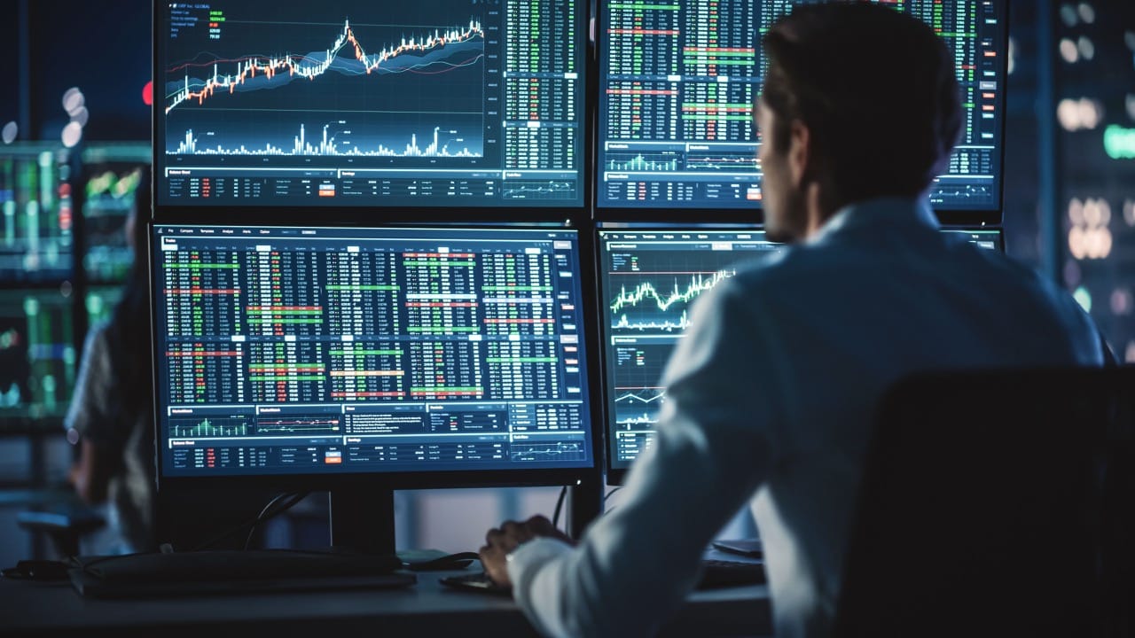 Person monitoring financial trading data on multiple screens showing stock charts and market statistics in dark room.