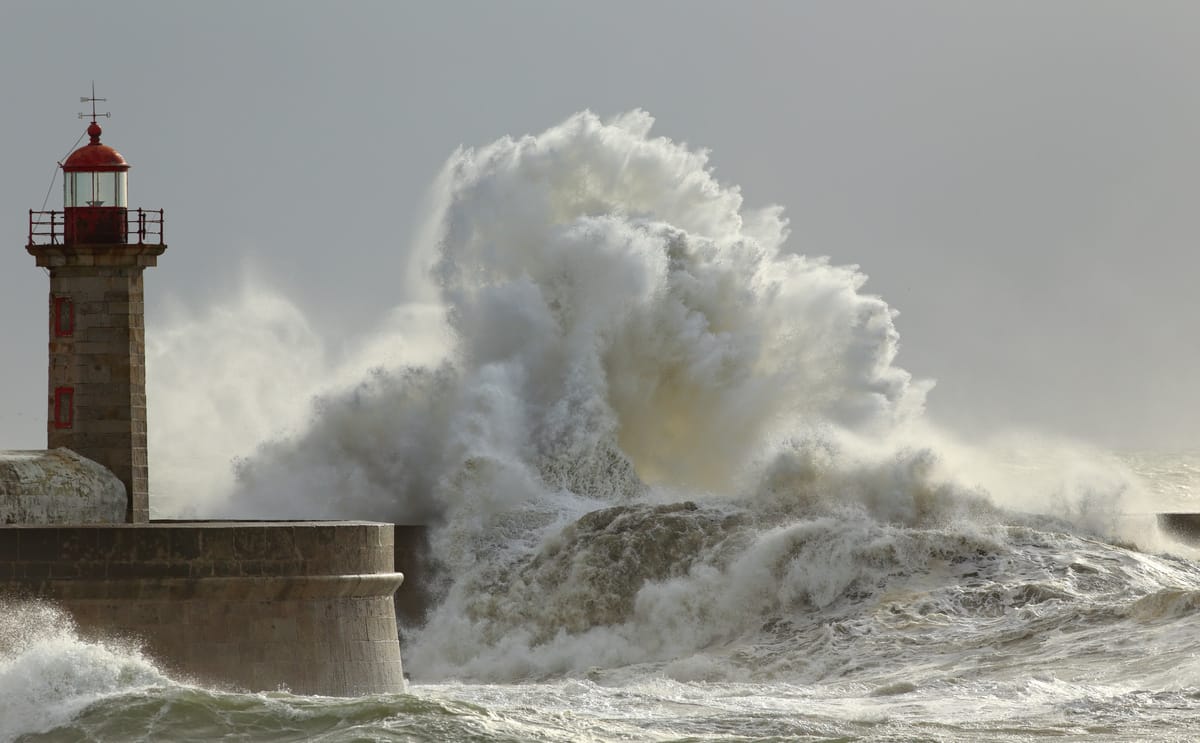 Massive wave crashes against harbour wall with red-topped lighthouse standing firm against stormy sea.