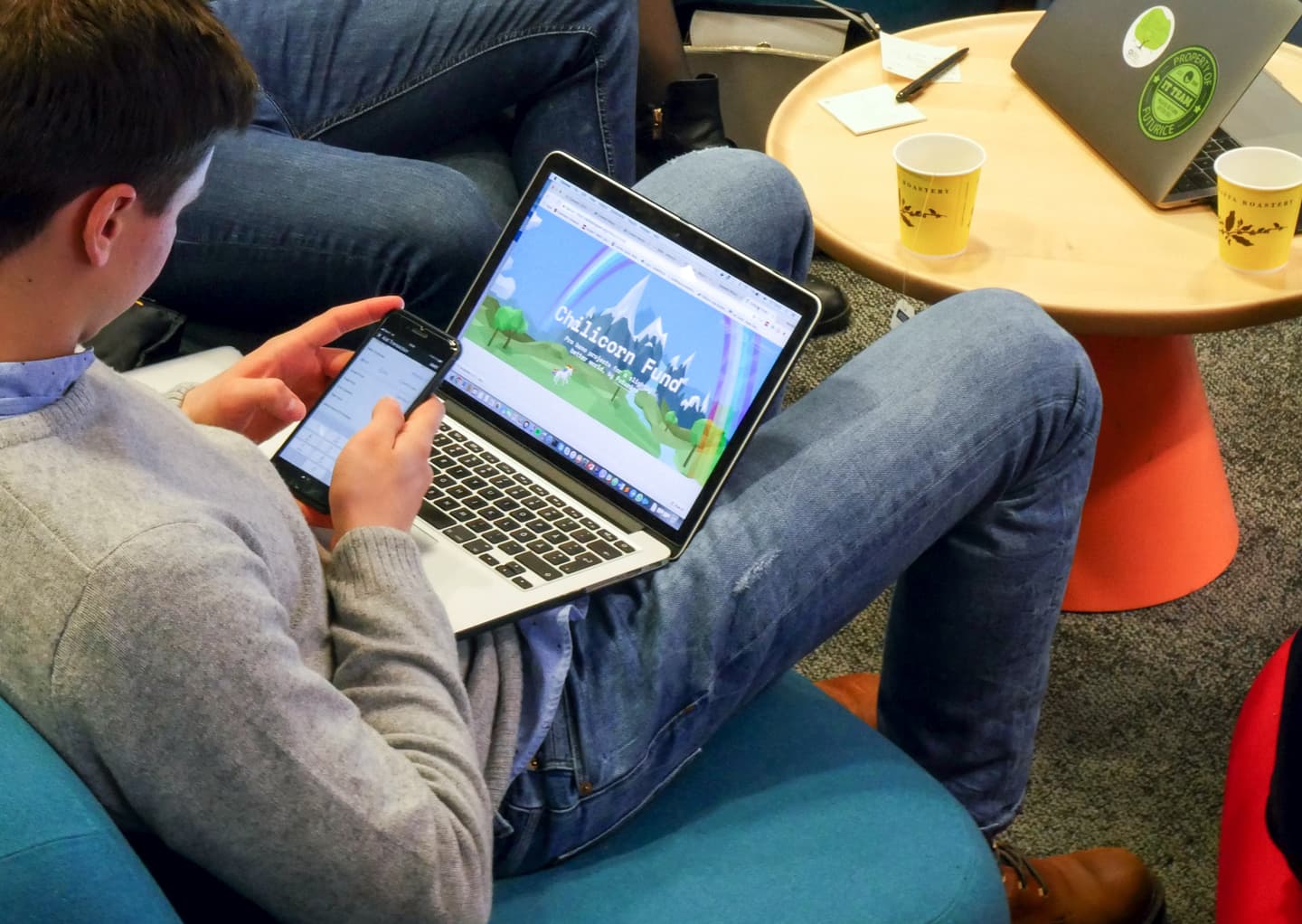 Person viewing "Unicorn Fund" website on laptop while checking phone, with coffee cups and another laptop on nearby table.
