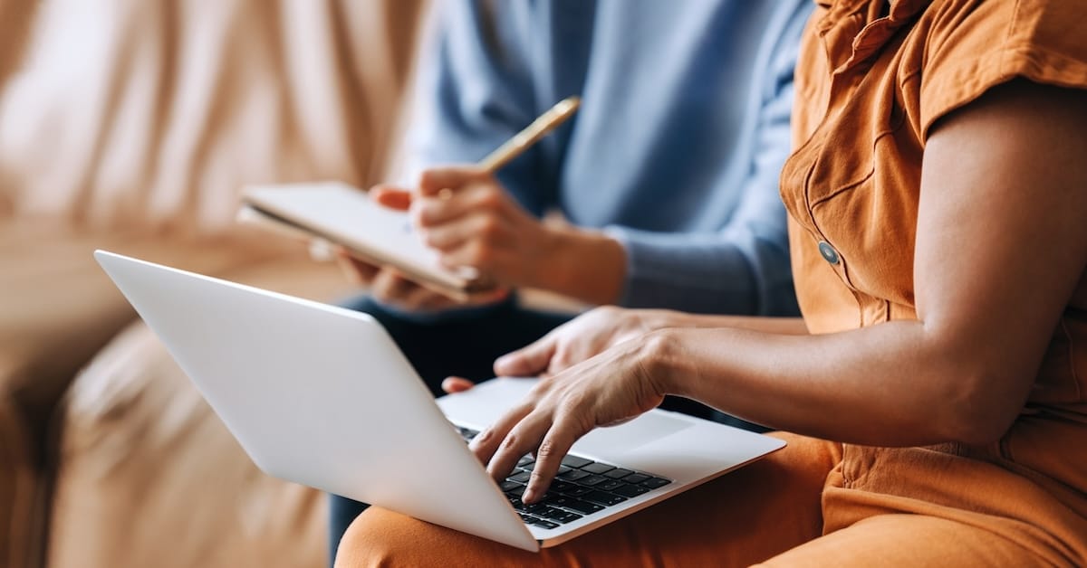 Person in orange outfit typing on laptop while another in blue shirt takes notes on a notepad during a meeting.