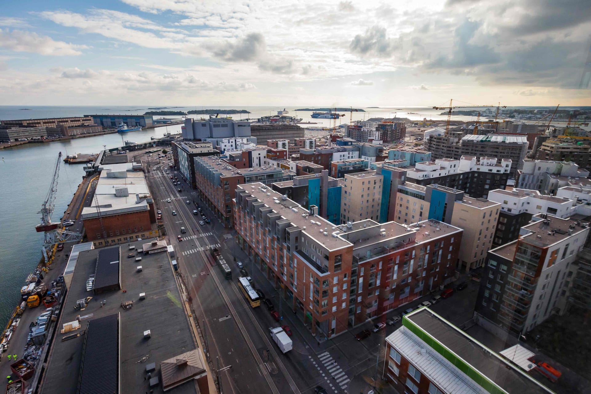 Aerial view of a coastal city with modern apartment buildings, harbour facilities and ships under a cloudy sky.