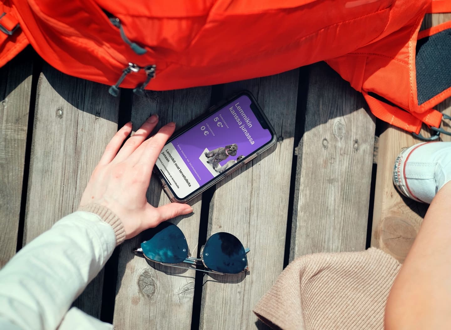 Hand using smartphone with purple app screen on wooden deck, next to orange backpack and sunglasses.