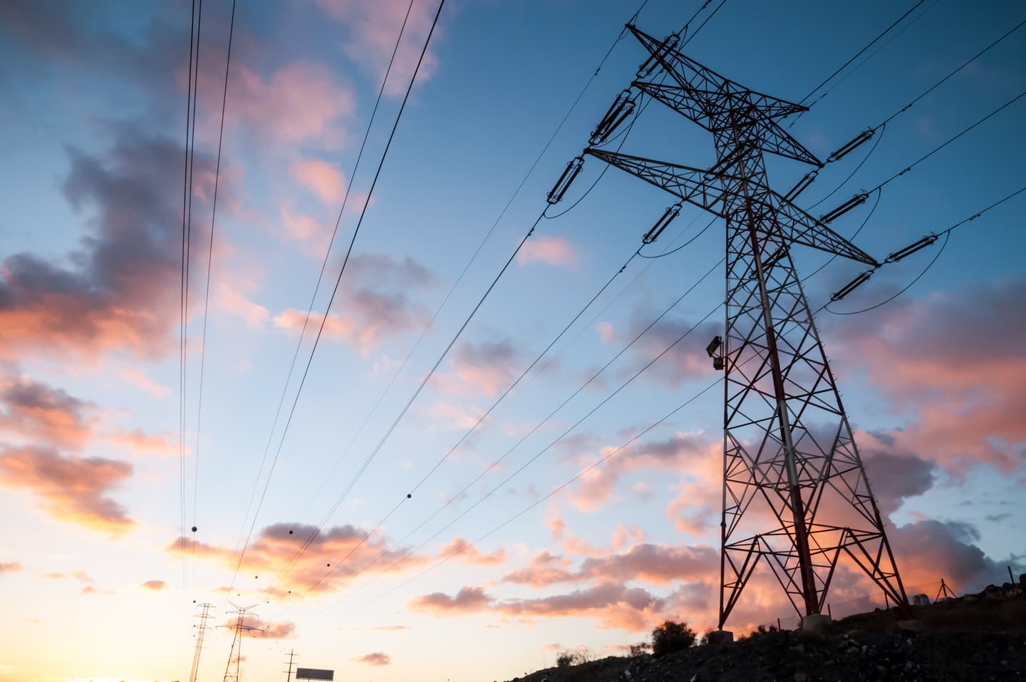 Electricity transmission tower silhouetted against a dramatic sunset sky with pink and orange clouds.