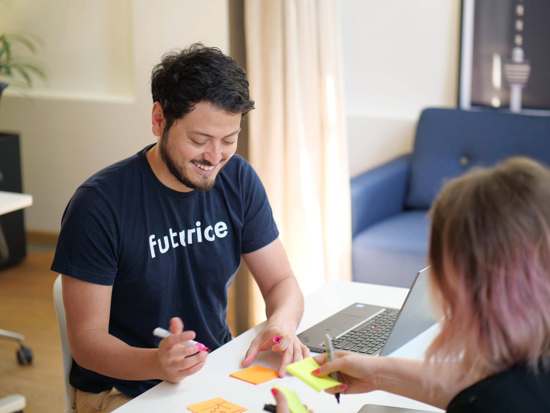 Person in navy "futurice" t-shirt smiling while collaborating with colleague using colourful sticky notes and laptop.