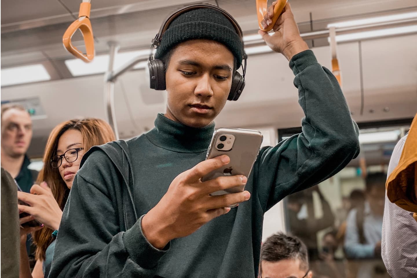 Person wearing headphones and beanie looking at smartphone while holding train handrail in crowded carriage.