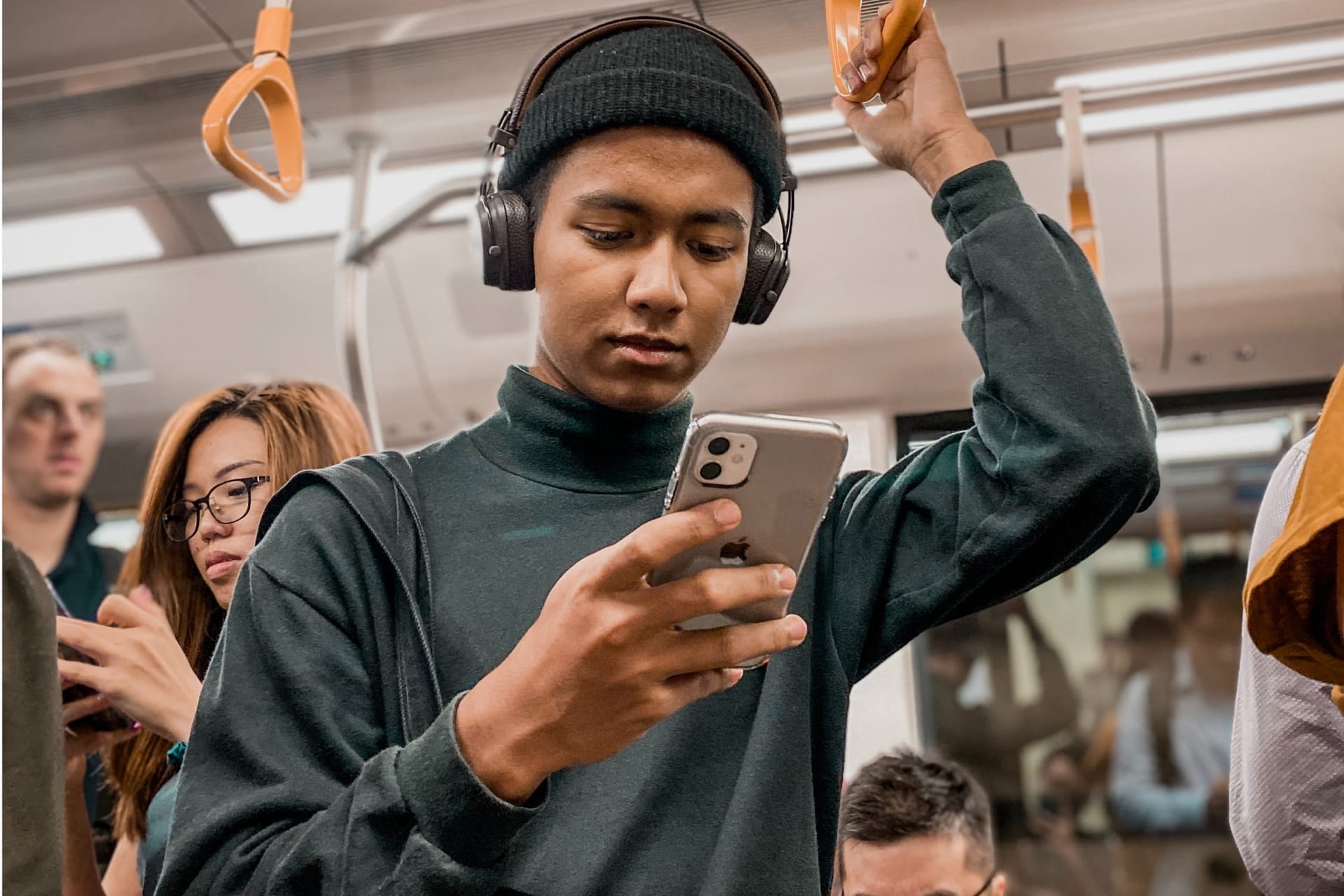 Person wearing headphones and beanie looking at smartphone while holding train handrail in crowded carriage.