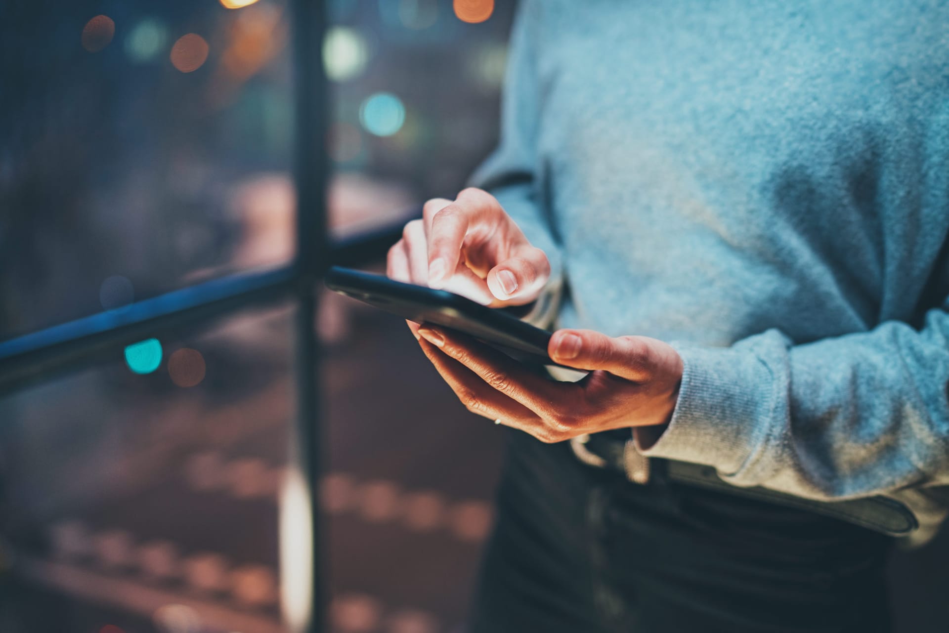 Person in blue sweater using smartphone at night with city lights blurred in background.