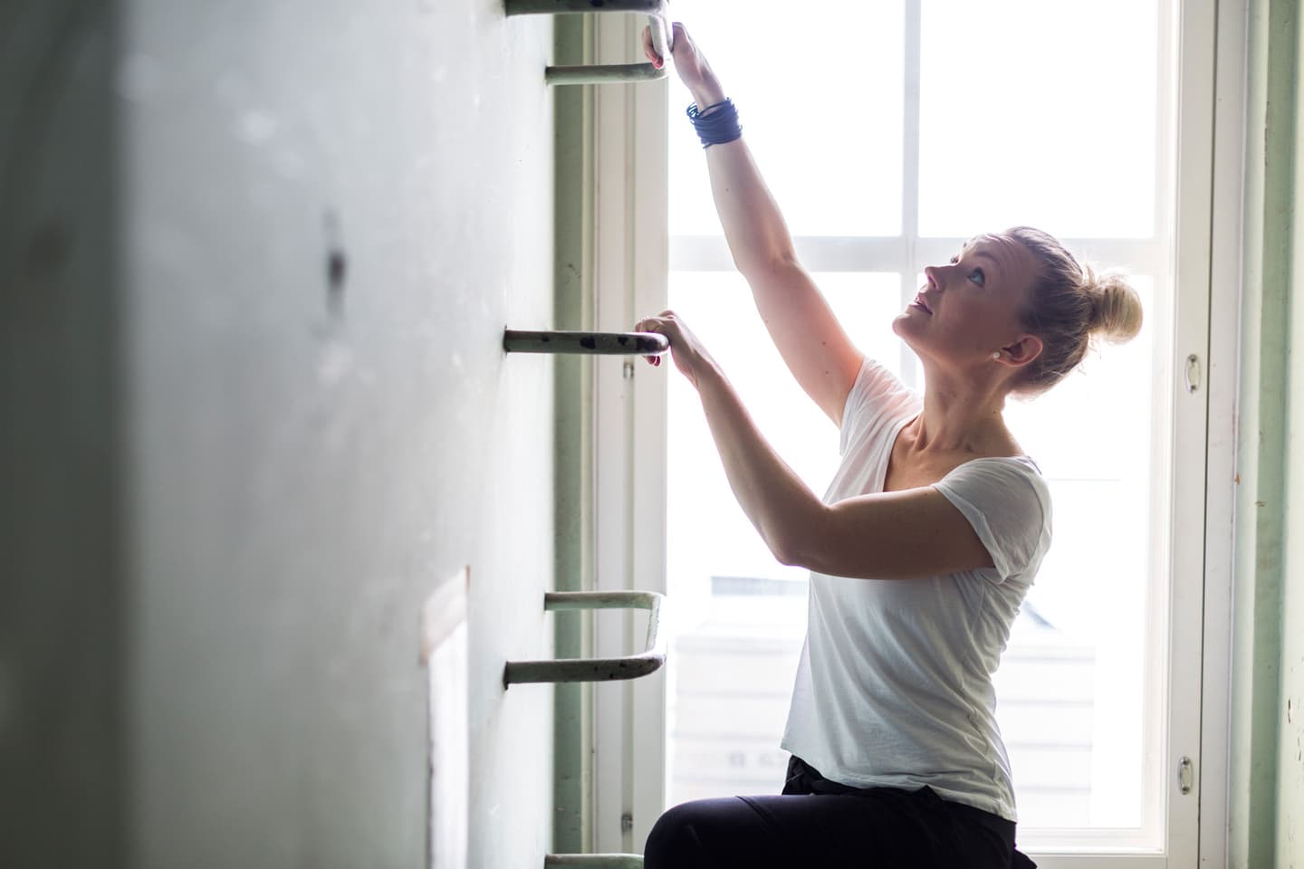 Woman in white top adjusting wall shelves near a bright window, wearing a blue bracelet.