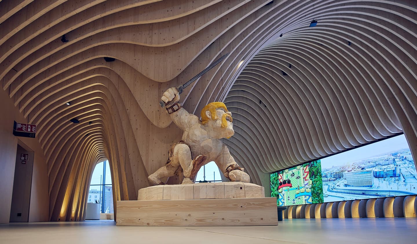 Large sculpture of a figure with golden hair wielding a sword beneath curved wooden ceiling in modern exhibition space.