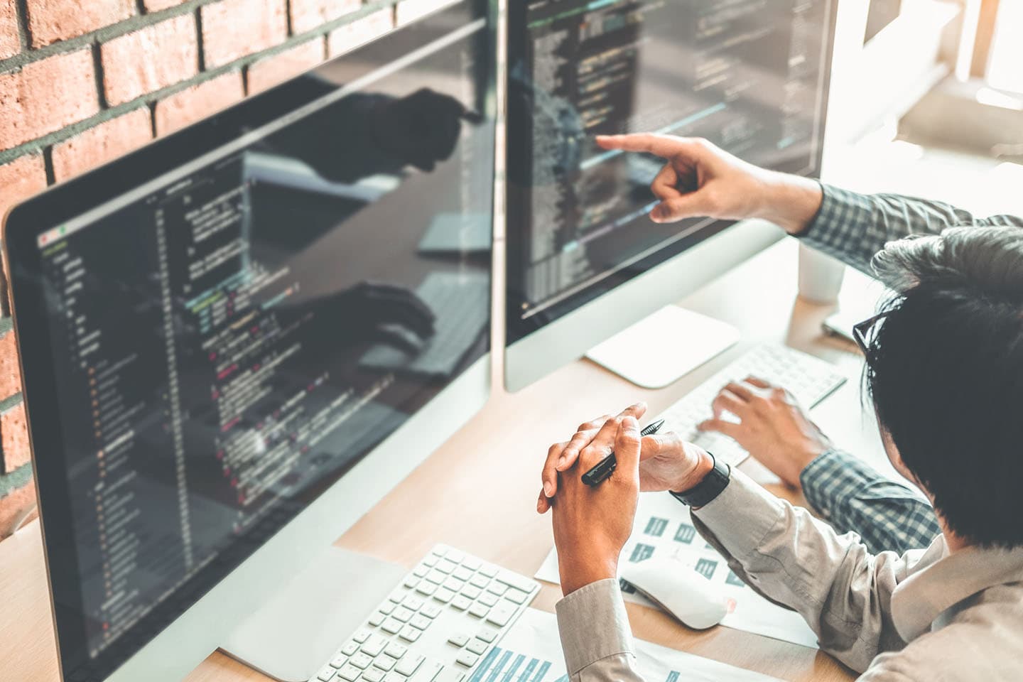 Programmer pointing at code on dual monitors while collaborating with colleague in office with brick wall background.