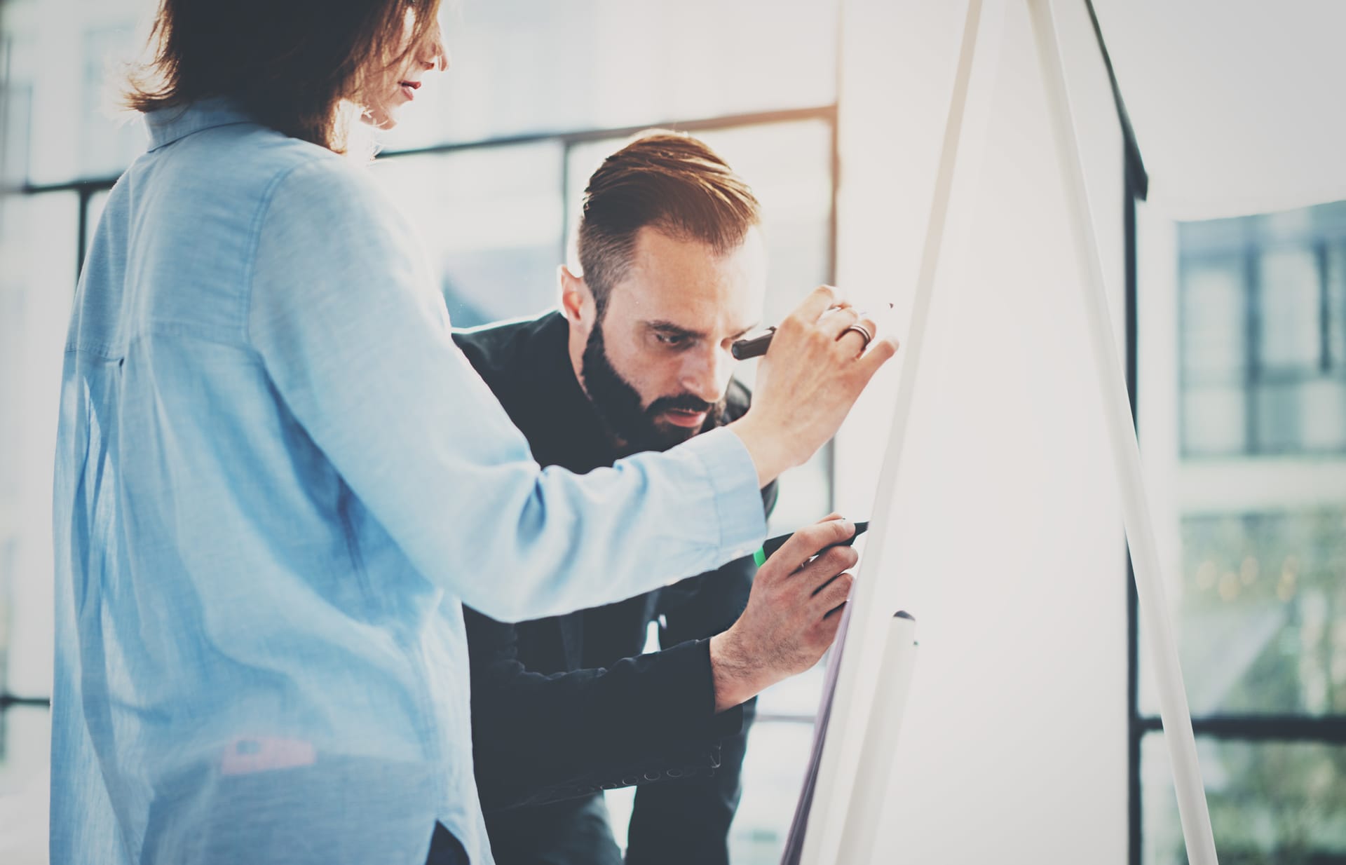 Two colleagues collaborating on a whiteboard, one in blue shirt and one in dark suit, in a bright office space.