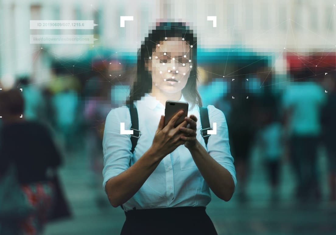 Person using smartphone with facial recognition technology overlay, digital identity markers visible in blue background.