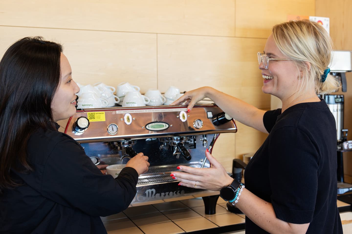 Two women in black tops smiling while operating an espresso machine in a café, with white cups stacked above.